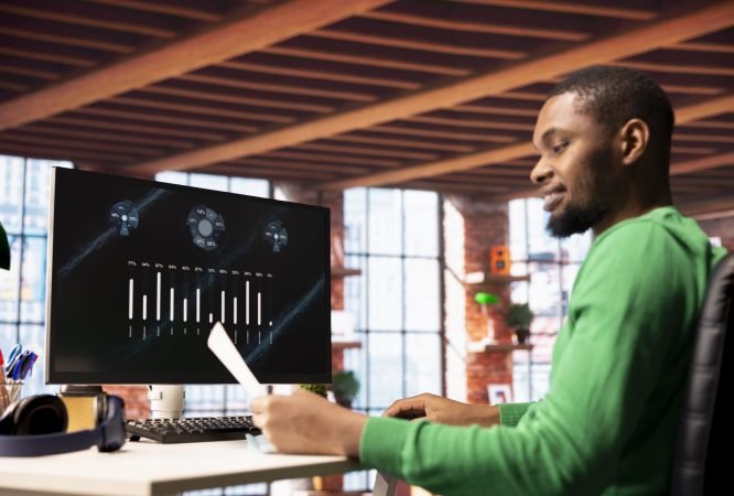 Smiling man in home office using financial application UI on PC screen showing company revenue and expenses. Accountant working from apartment doing annual profits review using computer display