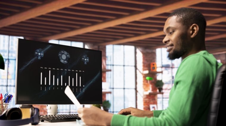 Smiling man in home office using financial application UI on PC screen showing company revenue and expenses. Accountant working from apartment doing annual profits review using computer display
